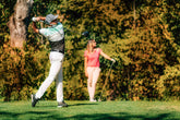 A couple at a golf lesson, with the man in the swing position and the woman observing, set in a lush golf course.