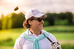 A smiling golfer with sunglasses and a cap, holding a golf club over his shoulder, enjoying a perfect golfing day.