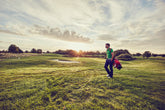 A golfer walking along the course at sunset, carrying a bag of clubs, with a scenic golf course in the background.