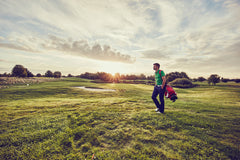 A golfer walking along the course at sunset, carrying a bag of clubs, with a scenic golf course in the background.