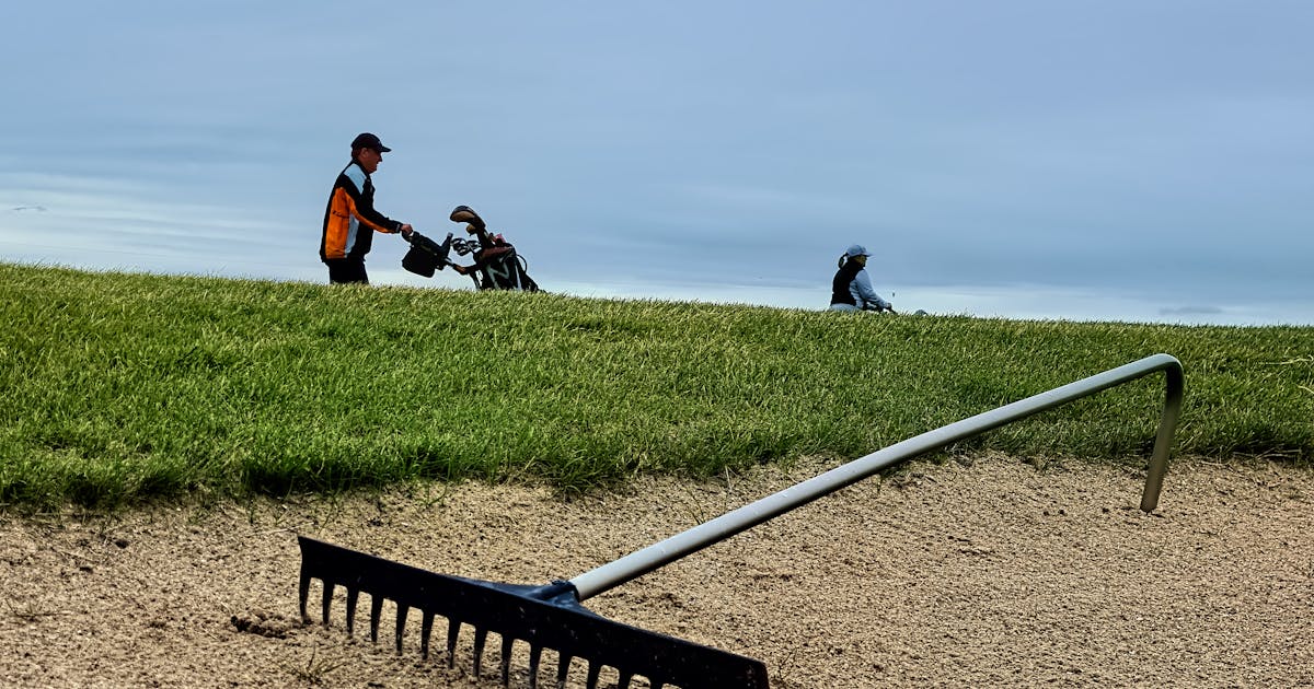 Two golfers walking on a course past a sand bunker under a cloudy sky