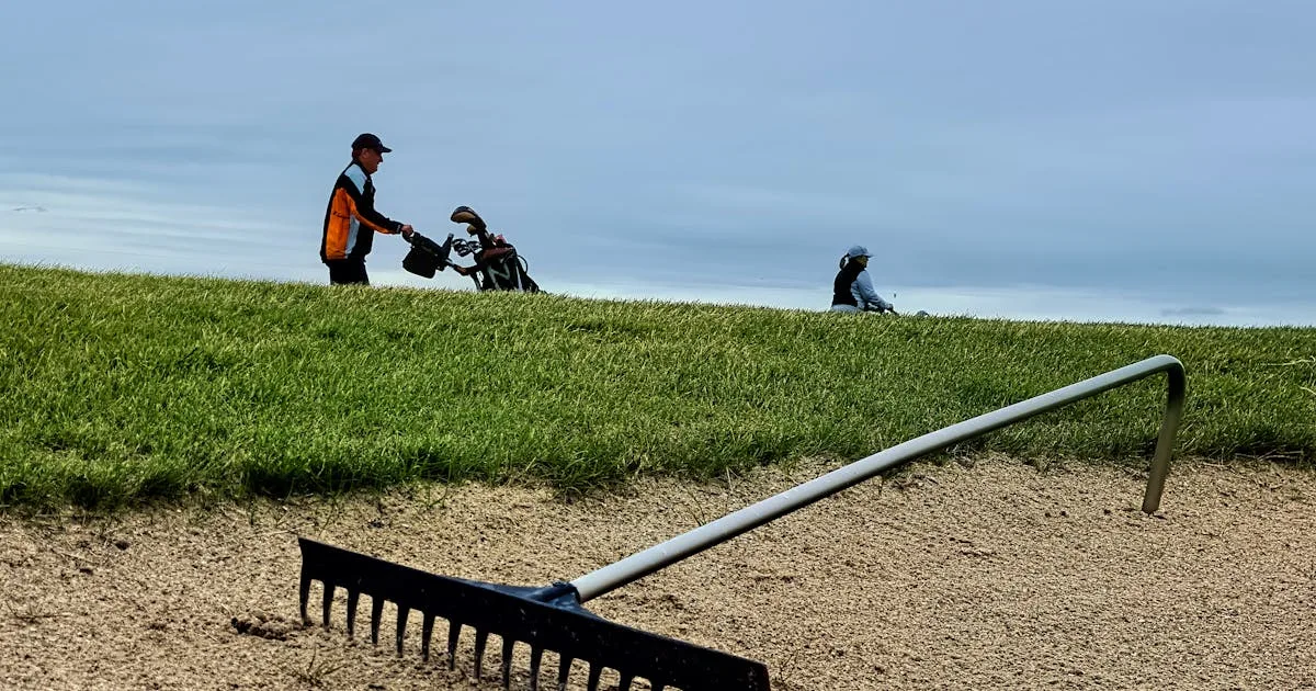Two golfers walking on a course past a sand bunker under a cloudy sky